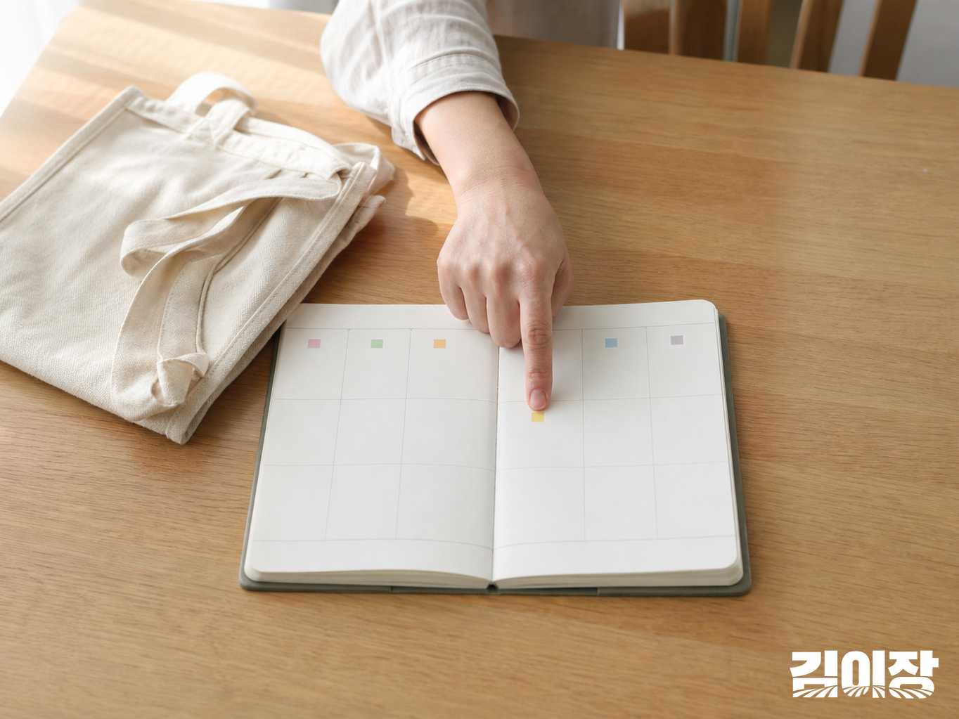 밝은 식탁에서 성인의 한 손이 색 표시가 있는 무문자 일정 수첩의 한 칸을 짚고, 옆에는 접힌 장바구니가 놓여 있어요.