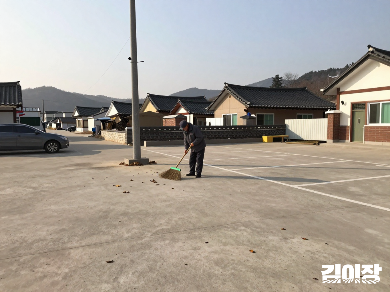 작은 마을 공동 주차장에서 주민 한 명이 긴 빗자루로 낙엽을 쓸고 있고, 주차 구획과 낮은 조명 기둥이 보이는 모습